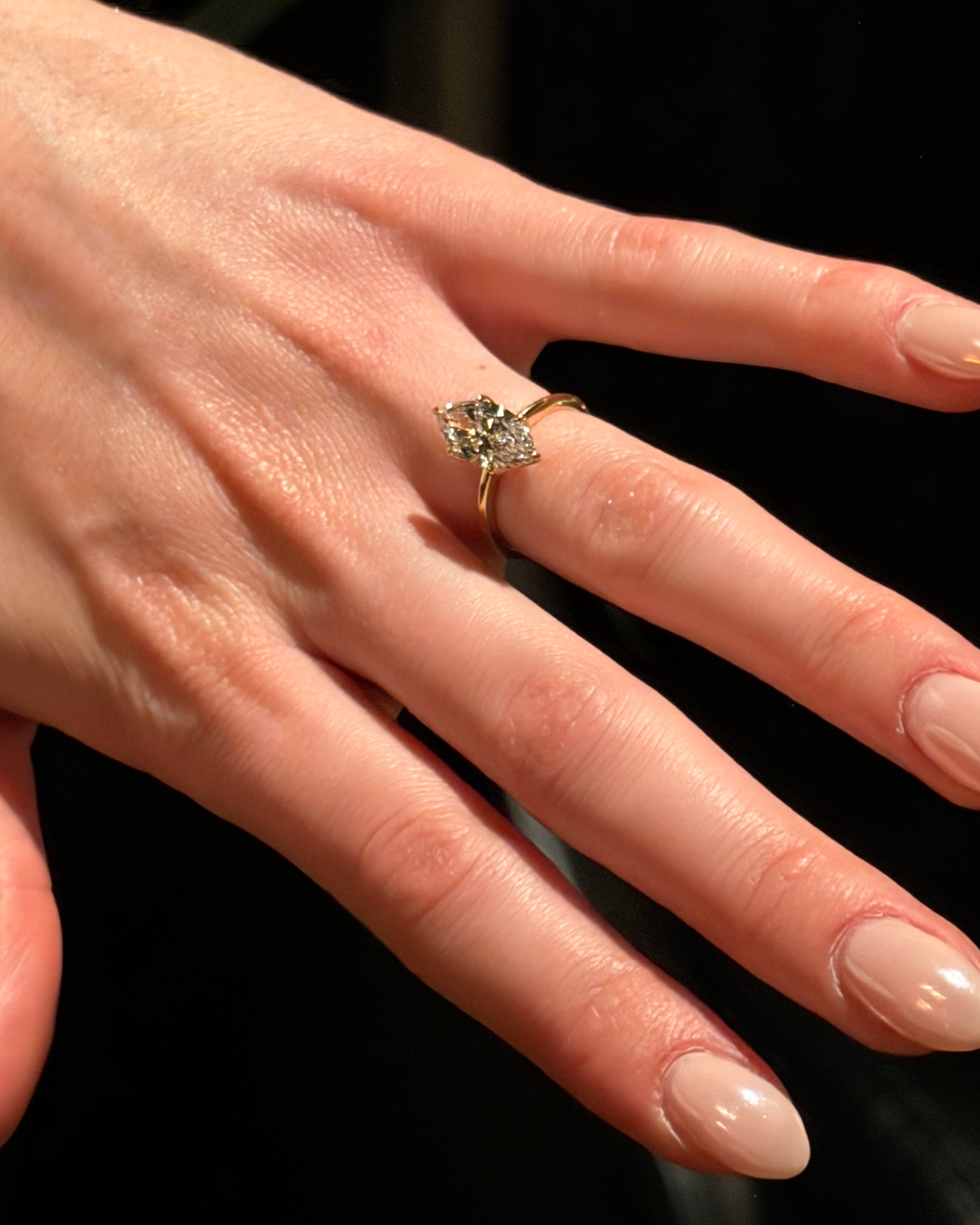 A close-up of a hand wearing a yellow gold ring with a marquise-cut lab grown diamond, set against a dark background.