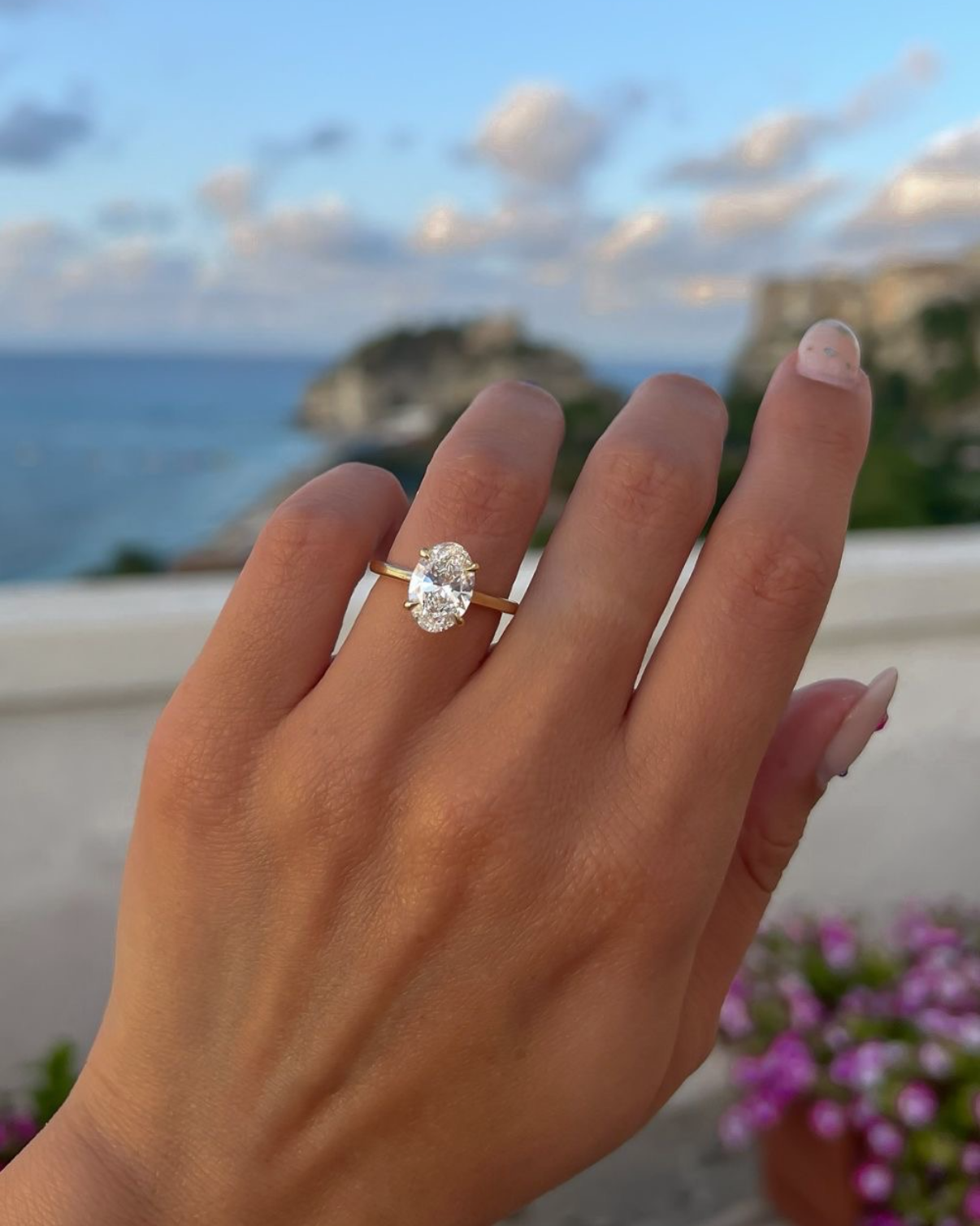 An engagement ring with a large oval diamond worn on a person's hand, set against an outdoor background of the sea and sky.